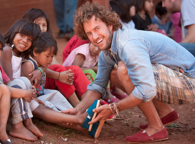 A man from TOMS helping children in need put on shoes, representing the brand's mission of giving back and resolving social challenges.