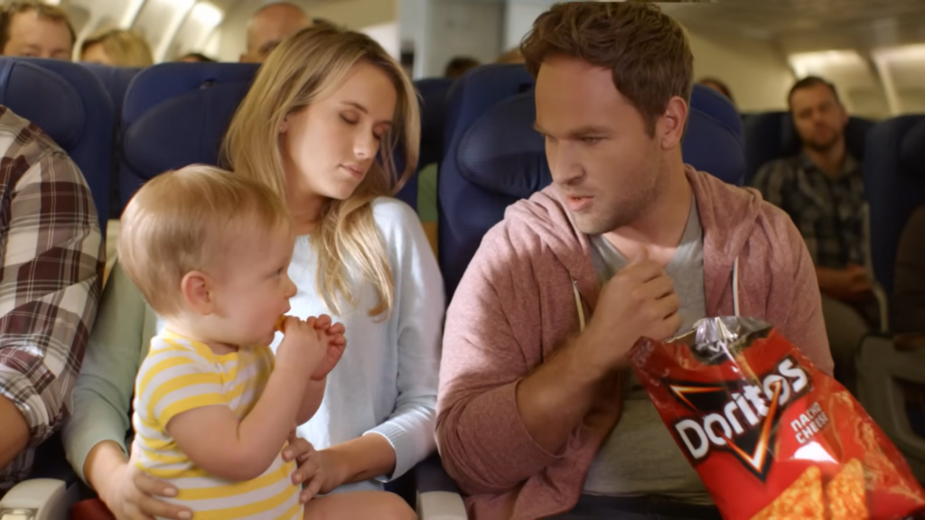 The protagonist in the 'Middle Seat' ad looks surprised and gestures towards the baby in the mother's lap, highlighting the unexpected twist