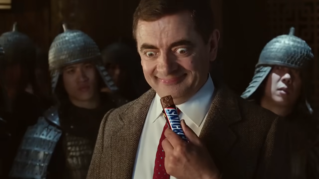 Mr. Bean holding a Snickers bar, preparing to take a bite.