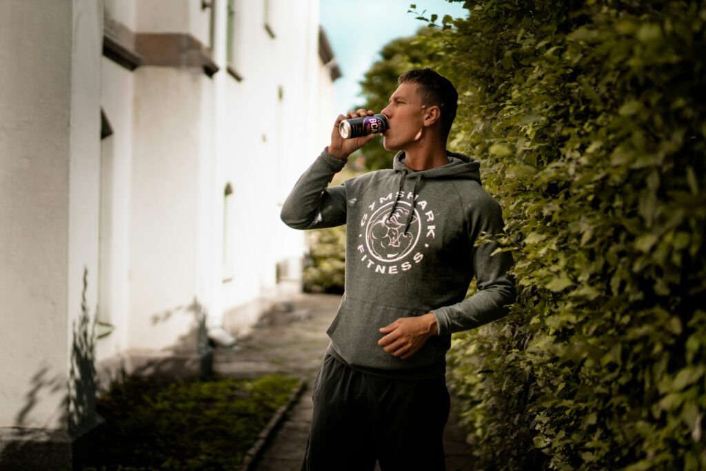 A man wearing a Gymshark hoodie, holding an energy drink and showcasing his influence by posting on social media in front of high-rise buildings.