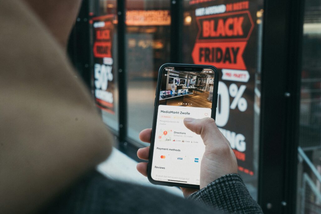 A person looking at their phone while standing in front of a store with a "Black Friday Sale" sign displayed prominently in the window.