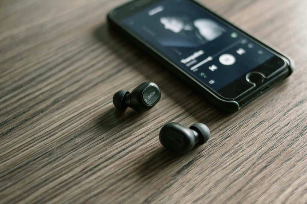 Two white wireless earbuds and a smartphone displaying the Spotify app on a wooden table.