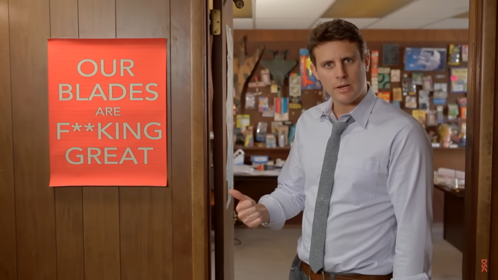 Michael Dubin pointing to the poster that reads "Our Blades Are F***ing Great" in the Dollar Shave Club advertisement.